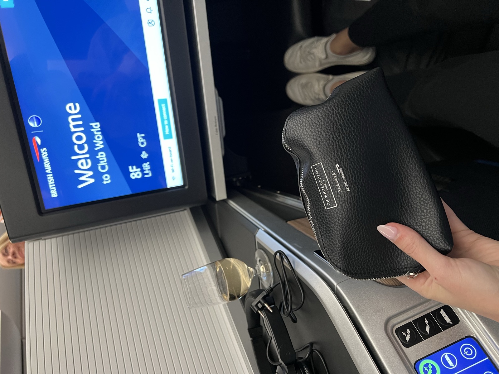 A passenger’s view of a British Airways Club World seat, with a welcome screen, glass of wine, amenity kit, and room to stretch out